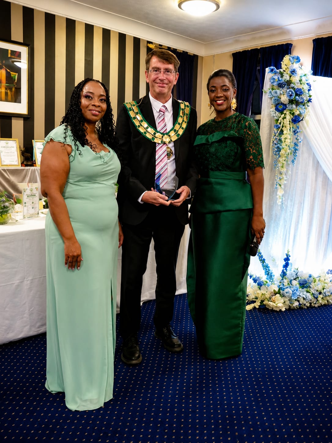 Lindona Panchoo and Founder & CEO Mrs Mariama C. Davies standing with Mayor David Rouane, who holds a recognition plaque.